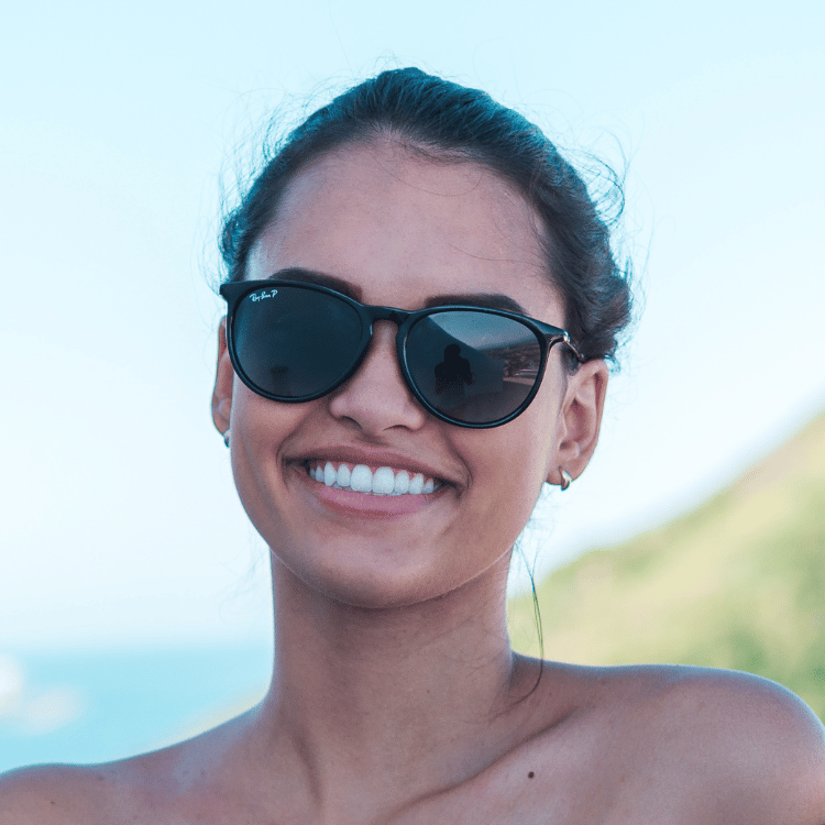 Picture of young women with dark hair pulled back off her face, wearing sunglasses with beach sceen in the background